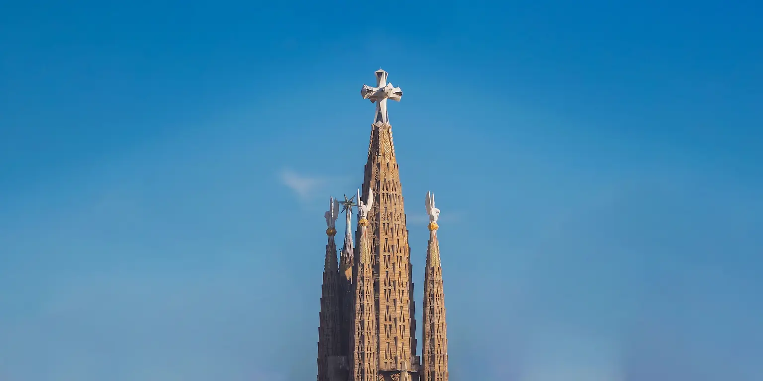 Vista vertical de las torres centrales de la Sagrada Familia destacando la torre principal coronada por una cruz sobre un cielo despejado.