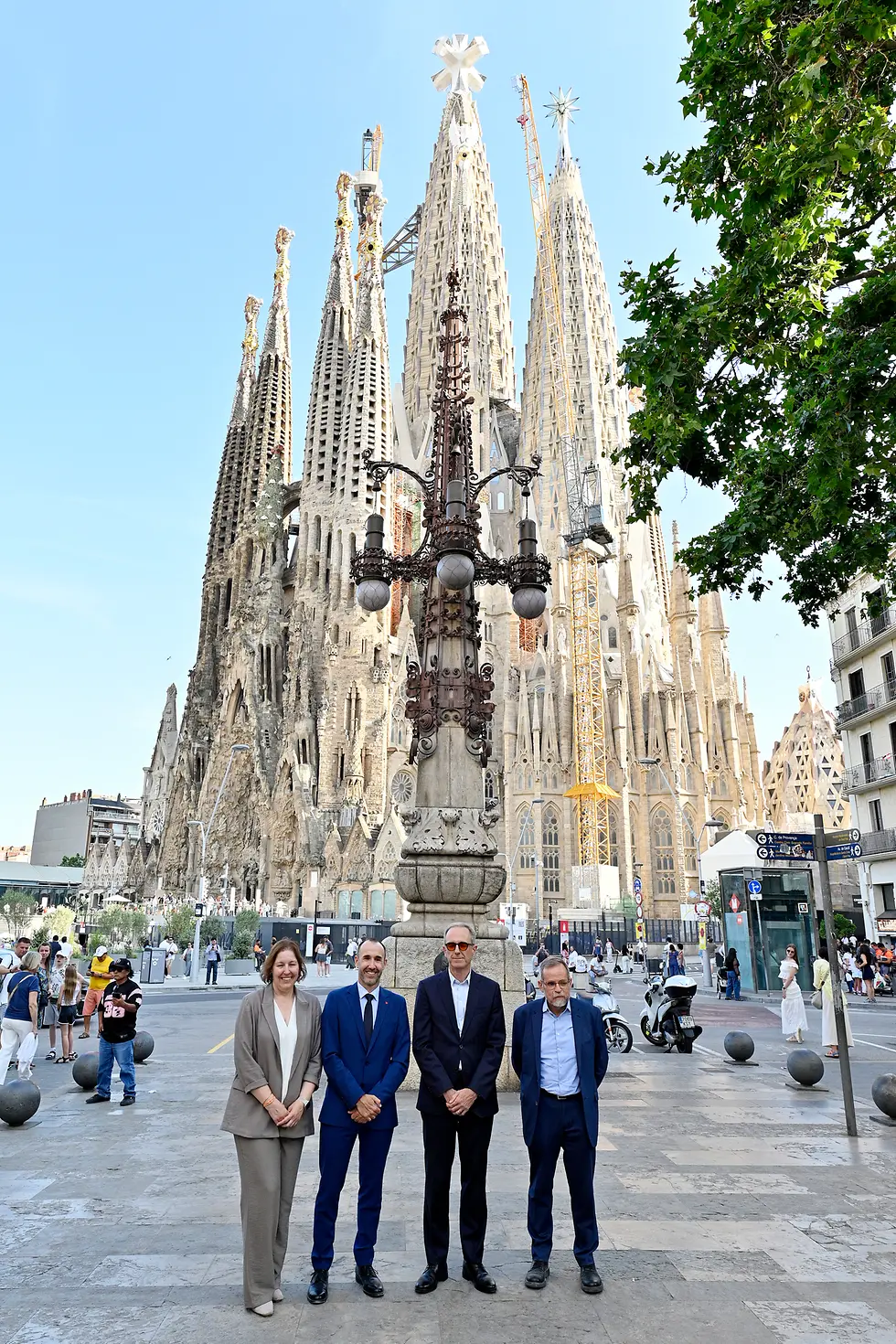 Vista general de la Sagrada Familia en Barcelona con sus torres centrales en construcción y cuatro personas posando en primer plano frente al templo en una plaza urbana con peatones y elementos de mobiliario urbano.