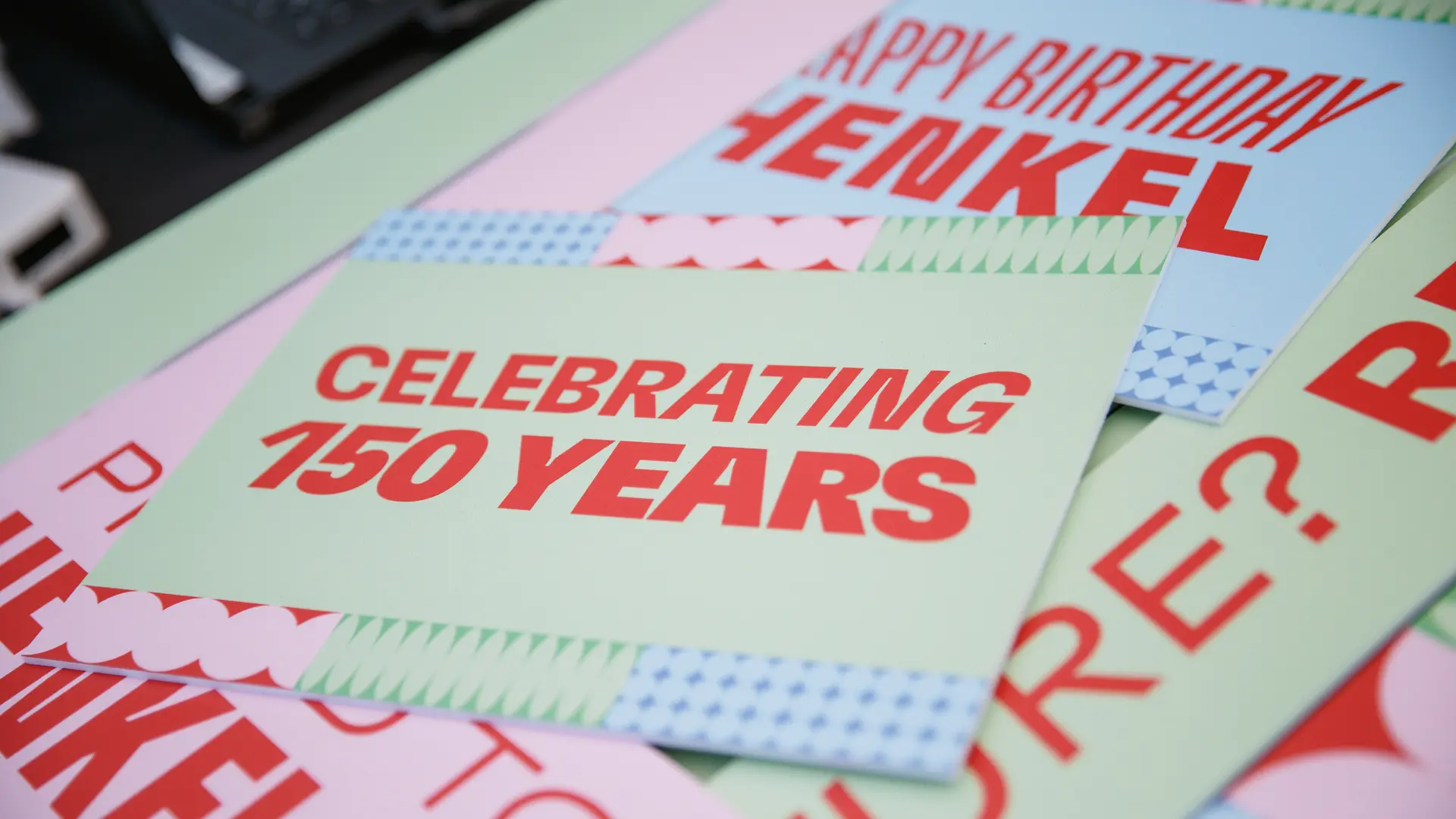 Material gráfico diseñado para la celebración del 150 aniversario de Henkel, con tarjetas con mensajes como “Celebrating 150 Years” y “Happy Birthday Henkel”, sobre una superficie en tonos claros.