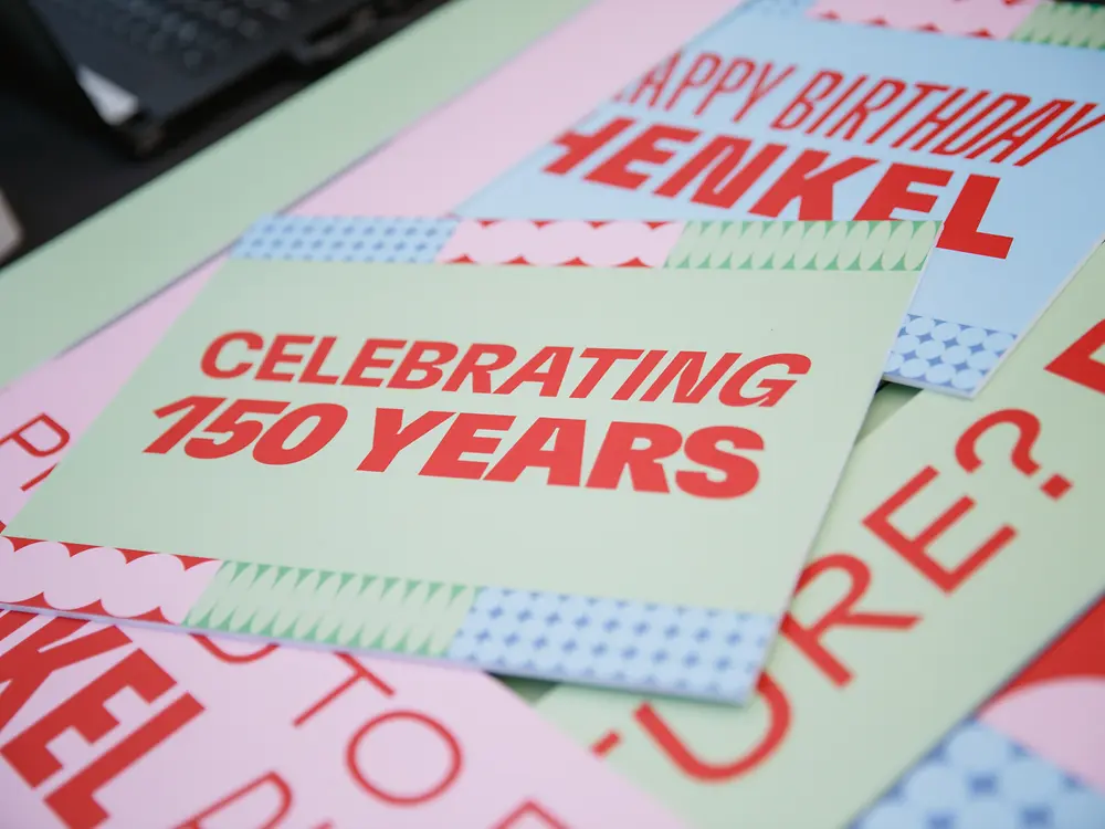 Material gráfico diseñado para la celebración del 150 aniversario de Henkel, con tarjetas con mensajes como “Celebrating 150 Years” y “Happy Birthday Henkel”, sobre una superficie en tonos claros.