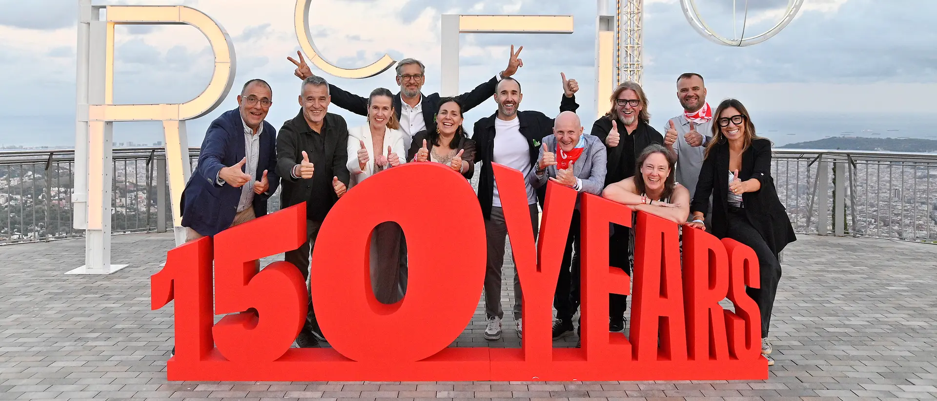 Integrantes del equipo directivo de Henkel Ibérica posan junto a una estructura con el mensaje “150 YEARS” durante el festival conmemorativo del 150 aniversario de Henkel, celebrado en Barcelona, con vistas panorámicas de la ciudad al fondo.