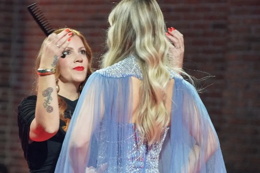 Profesional de peluquería trabaja sobre una modelo con cabello rubio de tonos múltiples y ondas suaves durante una presentación en pasarela.
