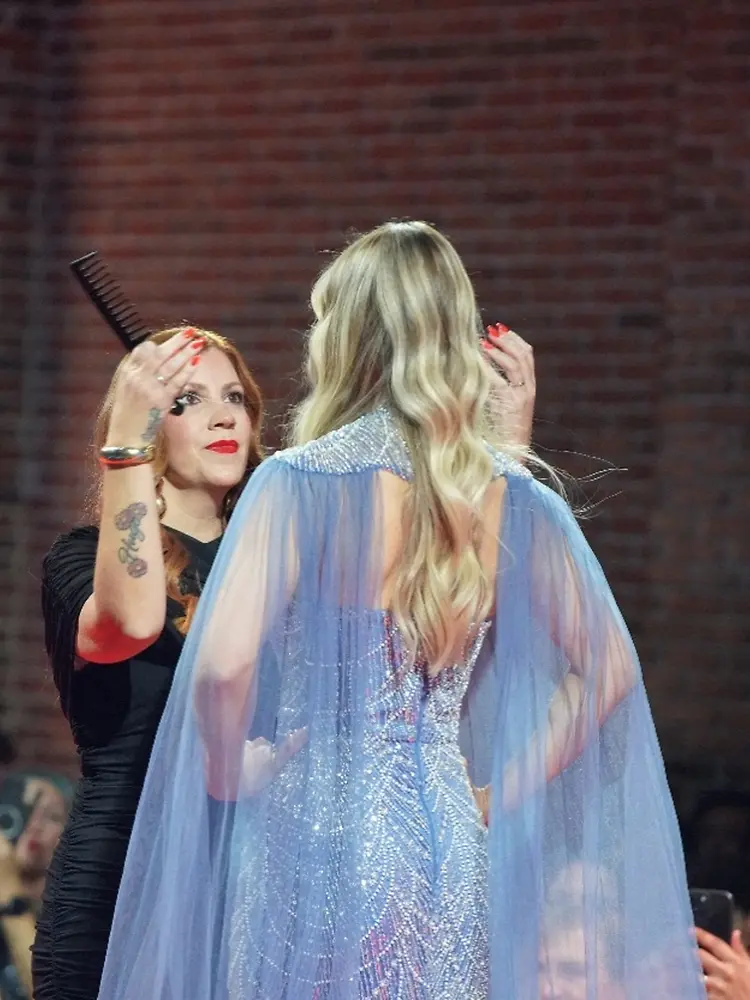 Profesional de peluquería trabaja sobre una modelo con cabello rubio de tonos múltiples y ondas suaves durante una presentación en pasarela.