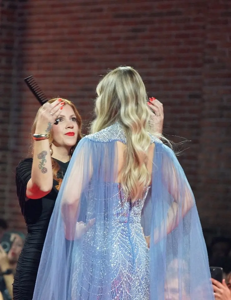 Profesional de peluquería trabaja sobre una modelo con cabello rubio de tonos múltiples y ondas suaves durante una presentación en pasarela.